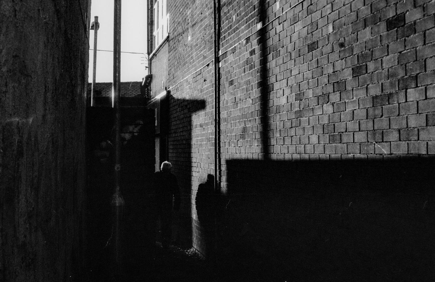 A man walks through a narrow alleyway between brick buildings, with strong shadows cast on the wall.