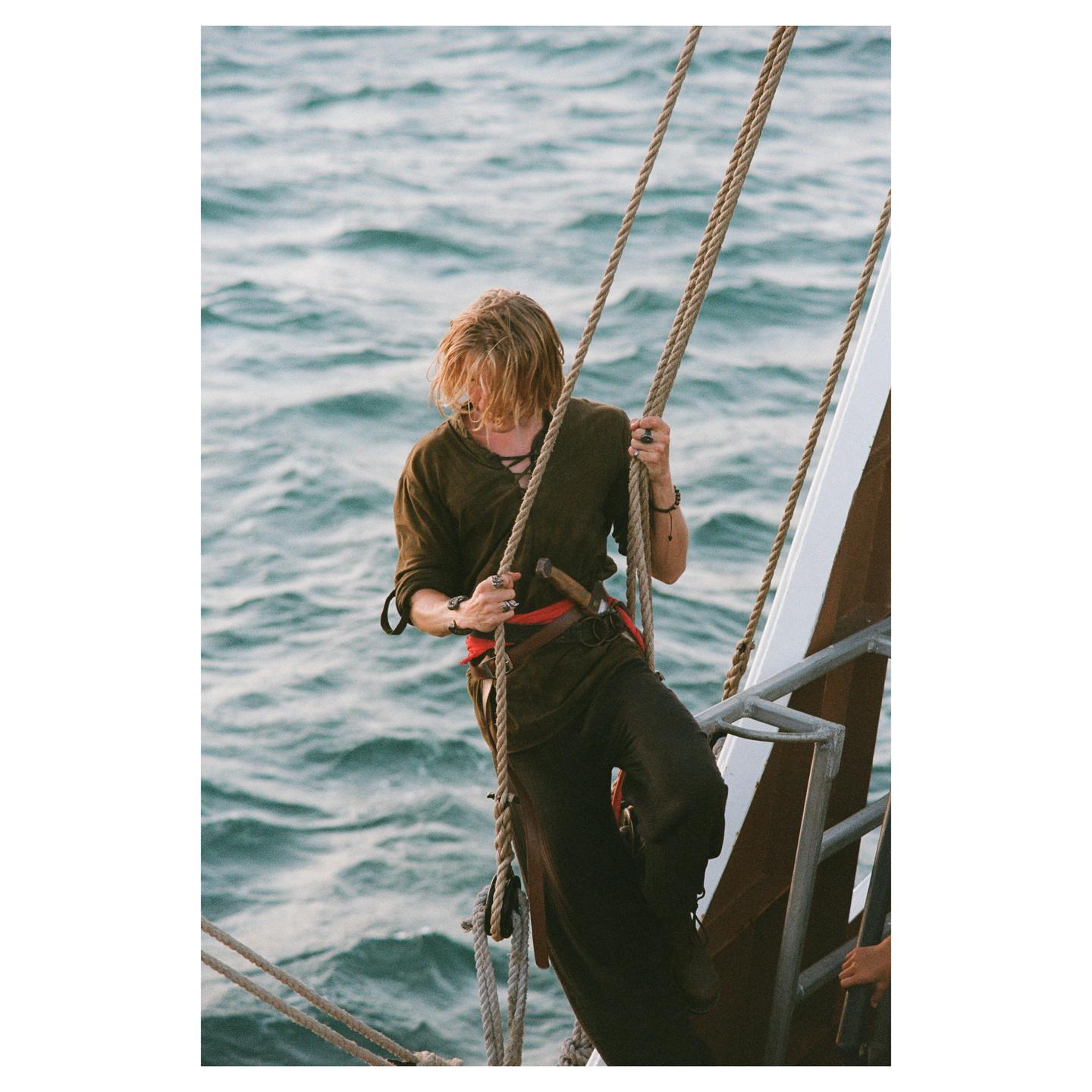 A man in period clothing holds onto ropes on a boat with the ocean in the background.