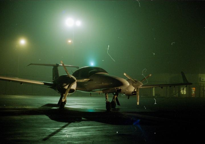 A small twin-engine propeller plane sits on an airfield at night, illuminated by bright lights.