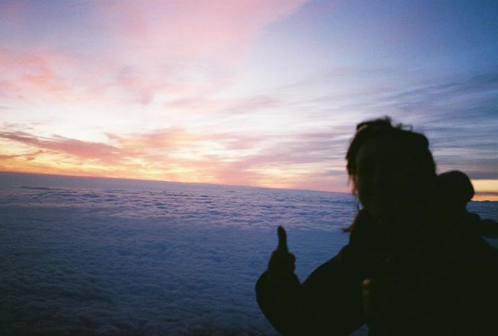 A person is silhouetted against a colorful sky above a sea of clouds, giving a thumbs up.