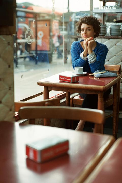 A woman with curly hair sits at a table in a cafe, looking towards the viewer.