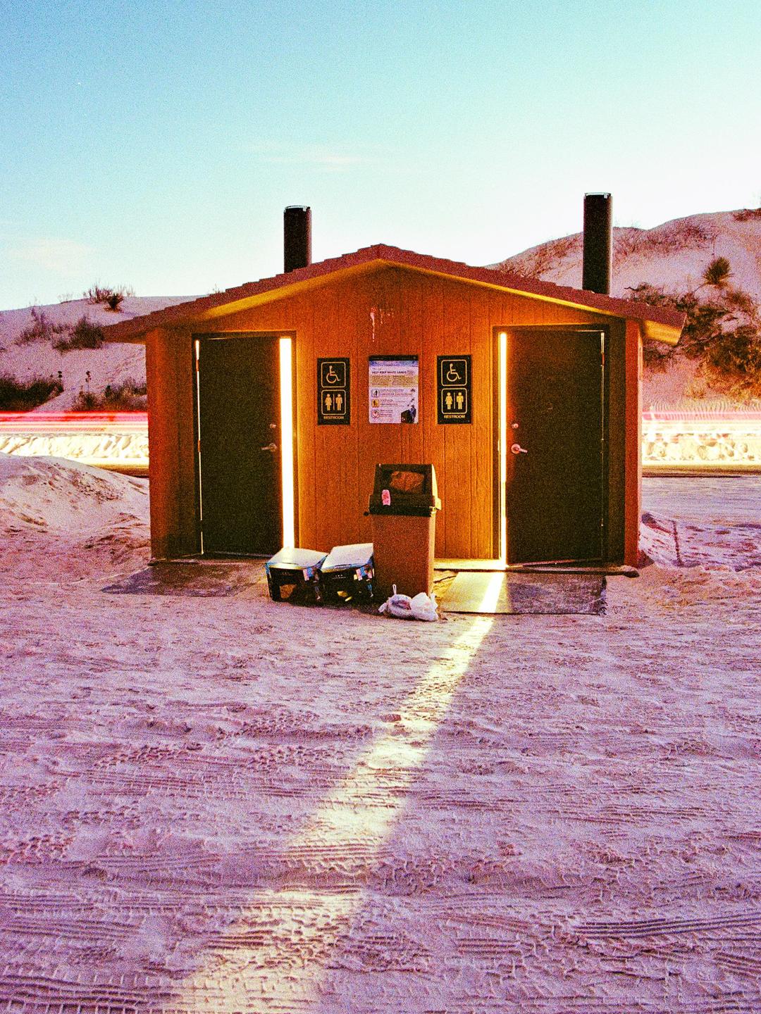 A small wooden building with two open doors and bright light inside stands on a sandy ground with tire tracks, with sand dunes in the background.