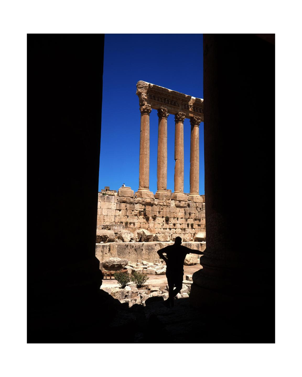 A silhouetted man stands in the shadow of a large column, looking out at ancient ruins with tall columns against a bright blue sky.