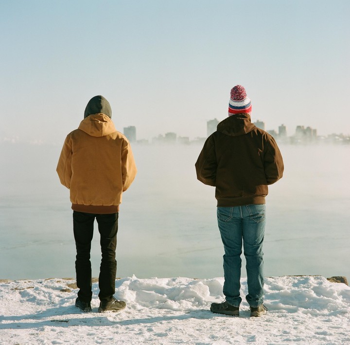 Two men stand on a snowy bank looking out at a foggy body of water with a city skyline in the distance.