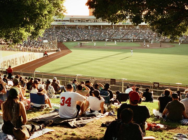 Spectators sit on a grassy hill watching a baseball game in a stadium.