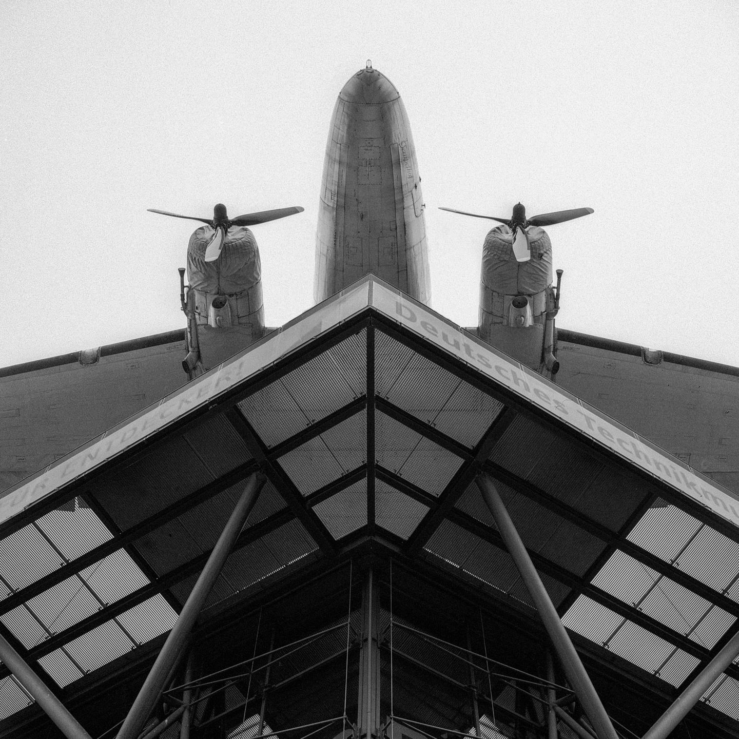 A low-angle view shows the nose and wings of a large airplane with two propellers, mounted above a modern building with a glass roof.