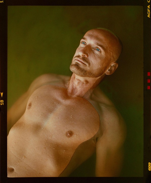 A man with blue eyes and a beard is partially submerged in water, looking upwards.