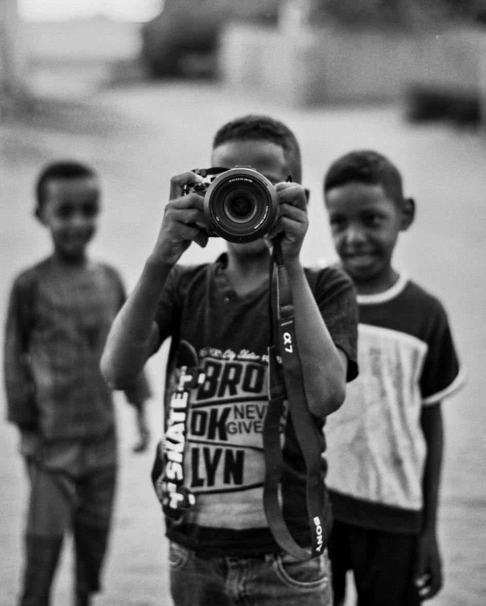 A boy in a "Brooklyn" t-shirt holds a camera to his face, with two other boys standing behind him.