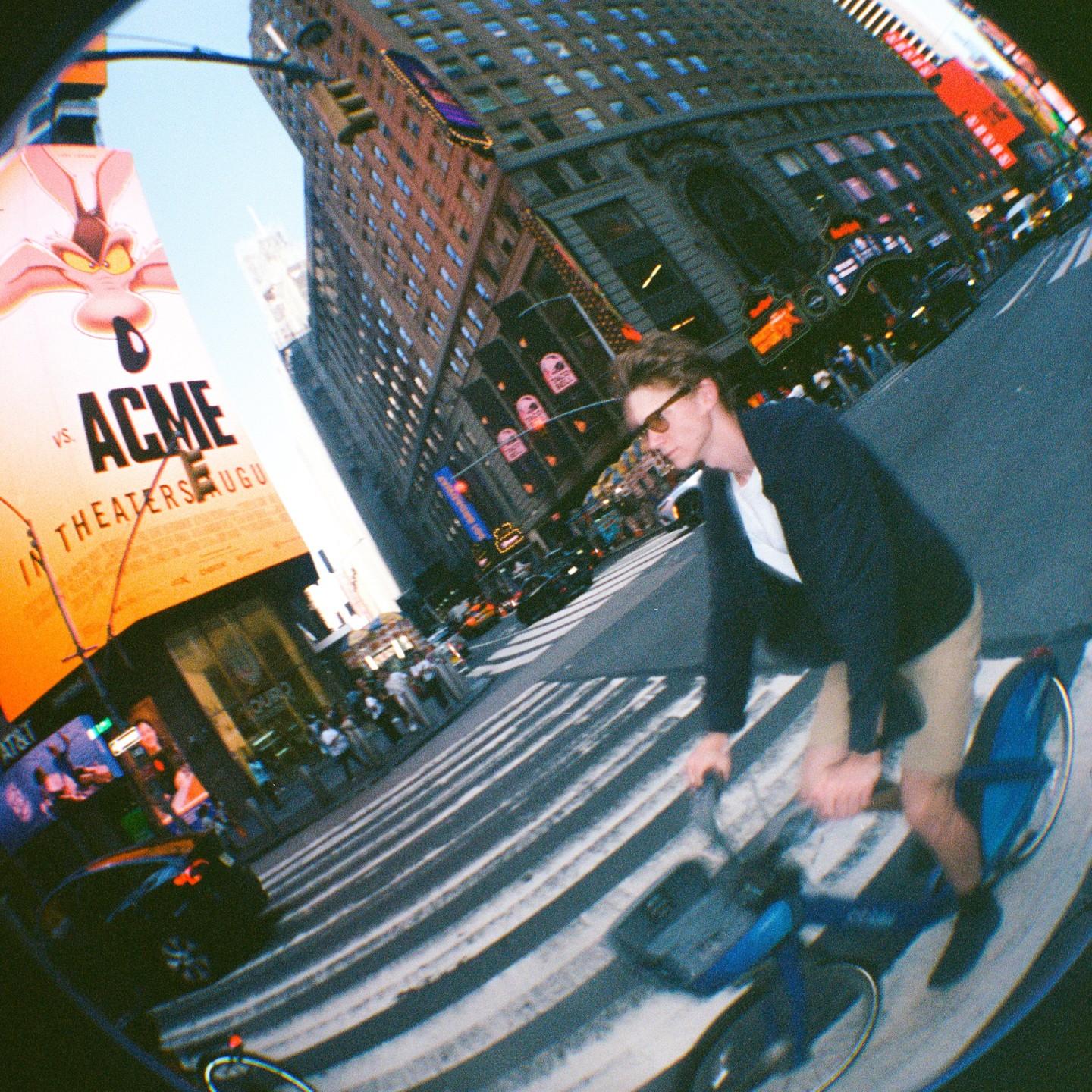 A man on a bicycle crosses a street in a city with tall buildings and a large billboard.