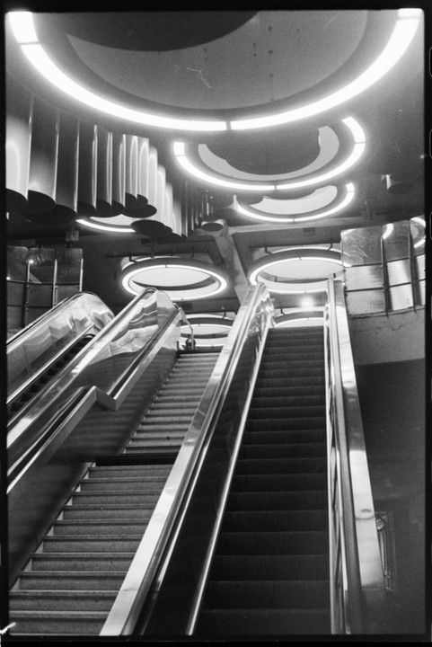 Two escalators and a staircase ascend towards a ceiling with circular and semicircular lights.