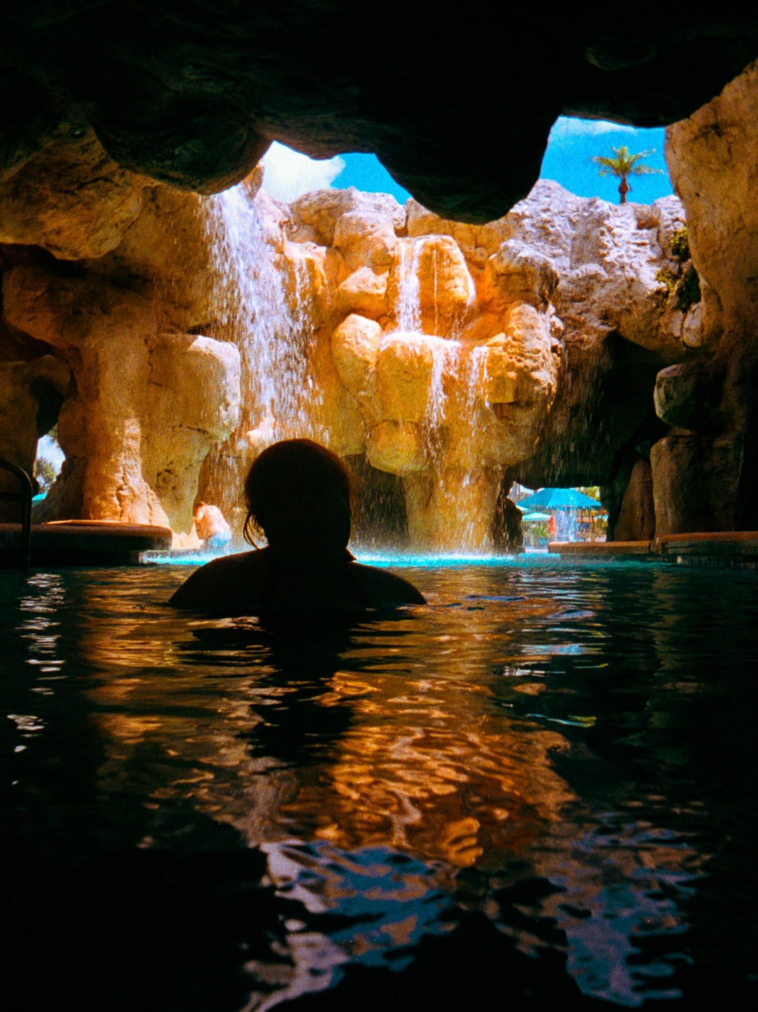 A person is in a pool looking at a waterfall cascading over rocks, with a palm tree visible in the distance.