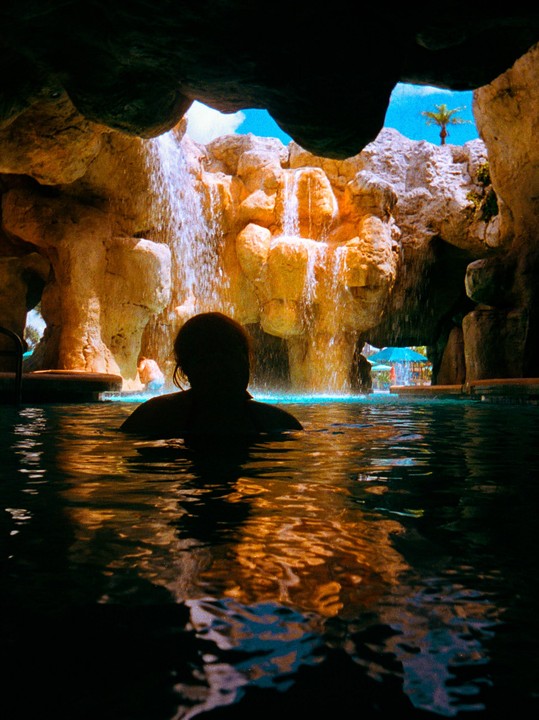 A person is in a pool looking at a waterfall cascading over rocks, with a palm tree visible in the distance.