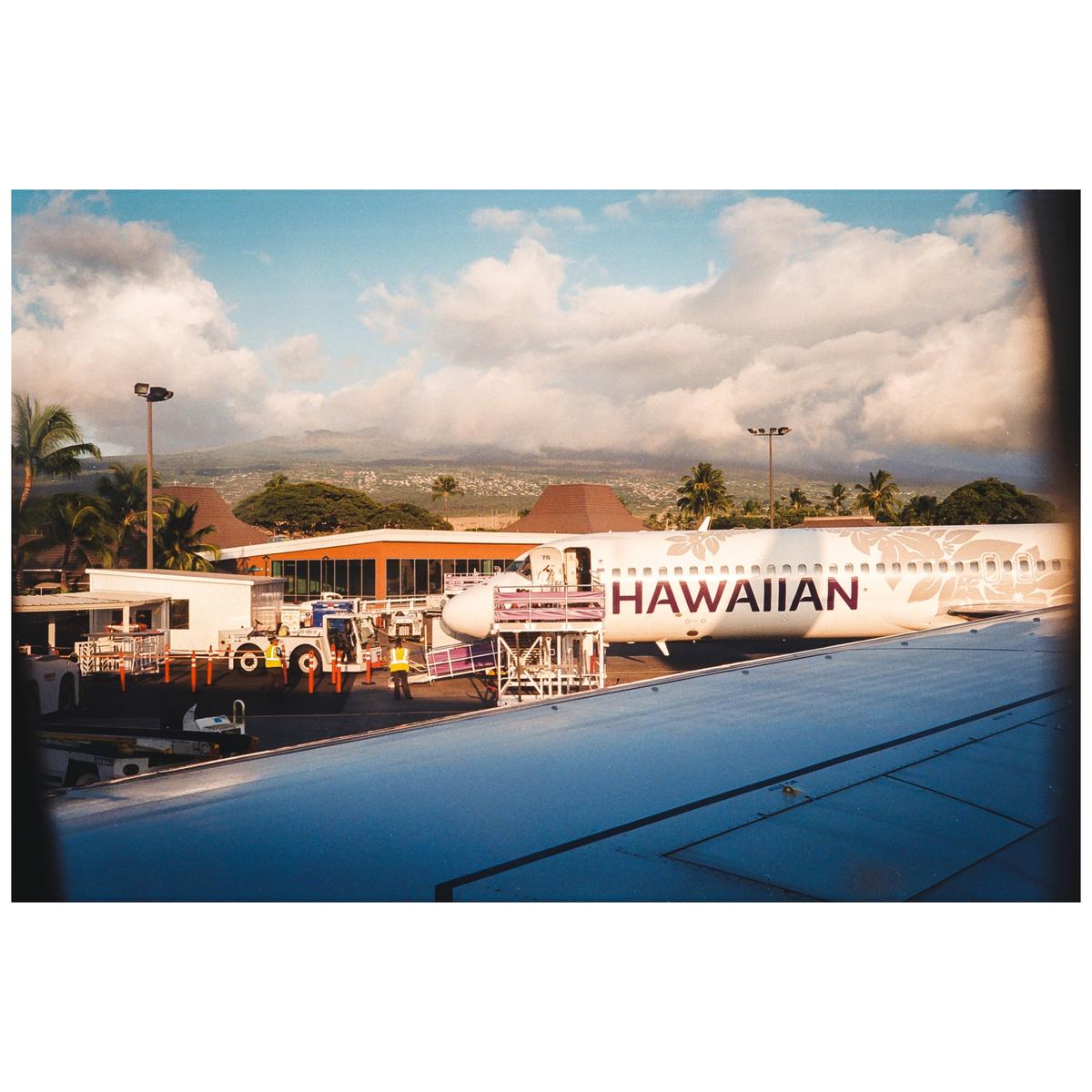 A Hawaiian Airlines airplane is parked at an airport gate with mountains in the background.