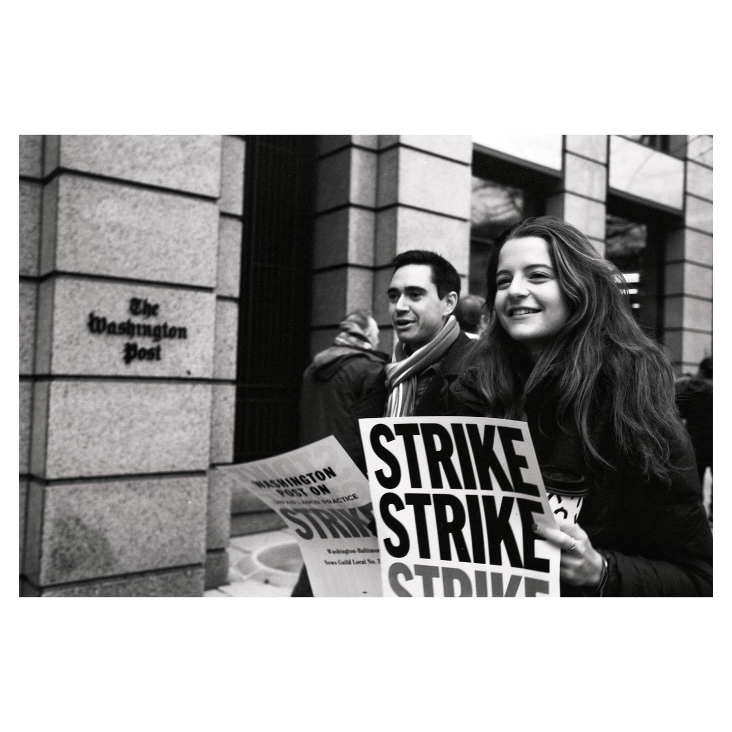 A woman and a man are holding "strike" signs in front of The Washington Post building.
