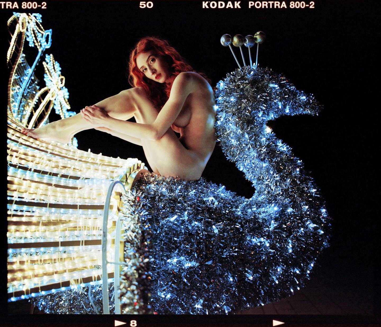 A nude woman with red hair sits on a large, illuminated peacock decoration.