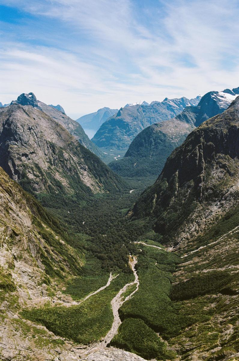 A deep valley with a river running through a forest is surrounded by mountains under a blue sky.