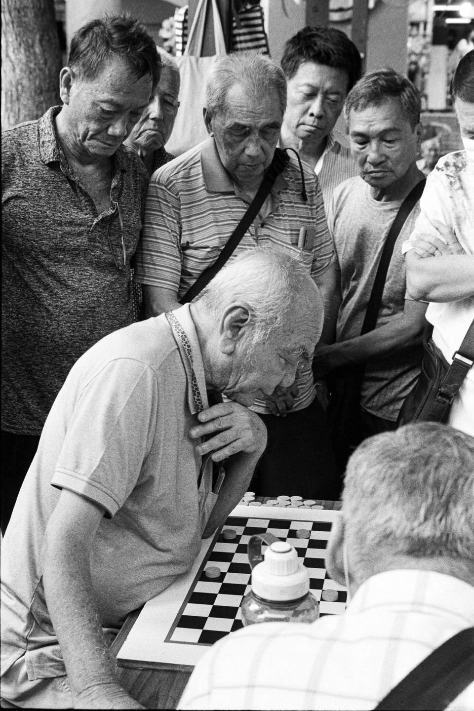 A group of men watches two other men play checkers on a board.