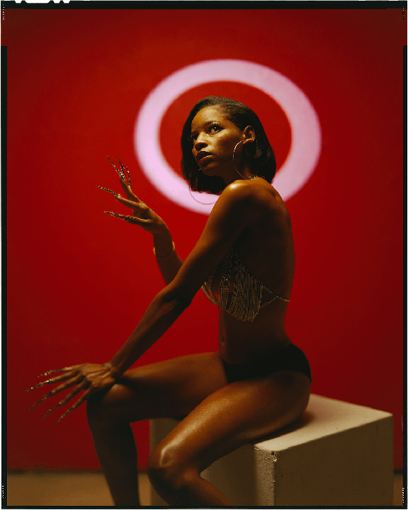 A woman with long fingernails and a chainmail top sits on a white block against a red background with a white circle.