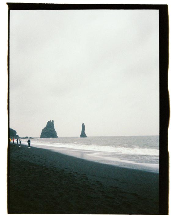 A group of people walk along a black sand beach with rock formations in the ocean under a cloudy sky.