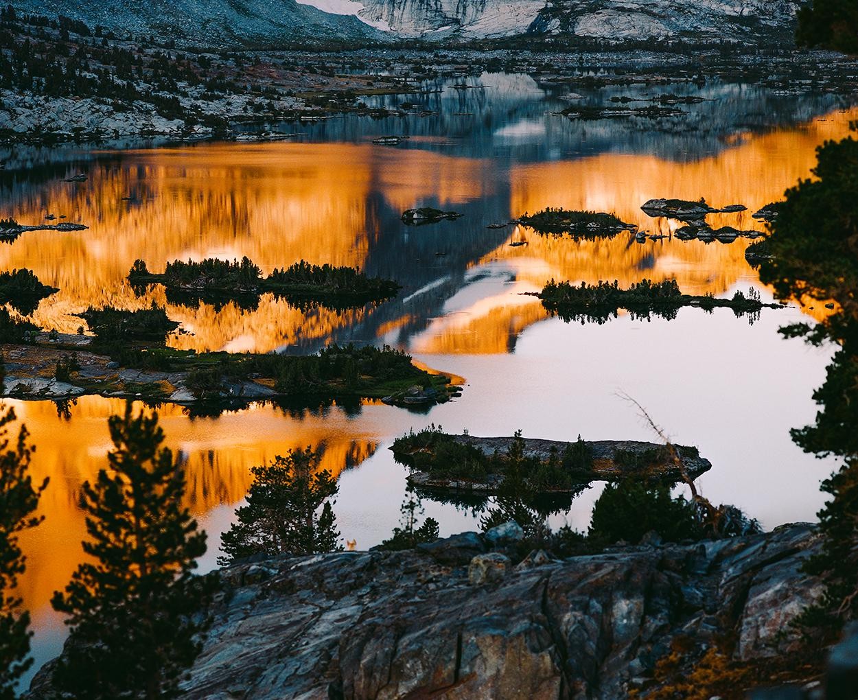 A lake with several small islands reflects the golden light of the sky and mountains.