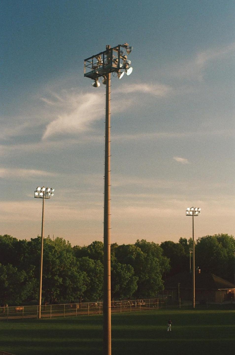 A baseball field with three tall light poles and a single outfielder is visible under a partly cloudy sky.