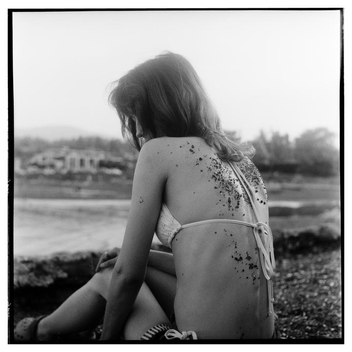 A woman in a bikini sits with her back to the viewer, covered in sand and small pebbles.