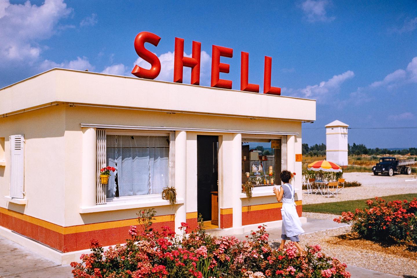 A woman walks past a Shell gas station with a large red sign on the roof, surrounded by blooming rose bushes.