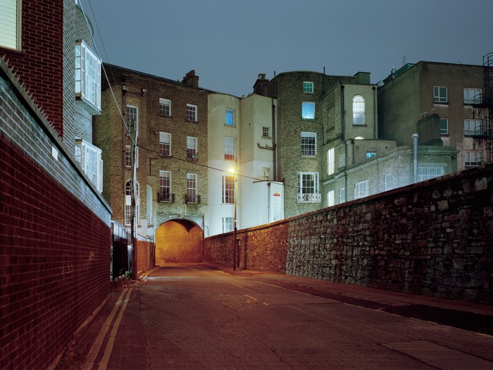 A narrow street at night is lined with brick buildings and a stone wall, leading to an illuminated archway.