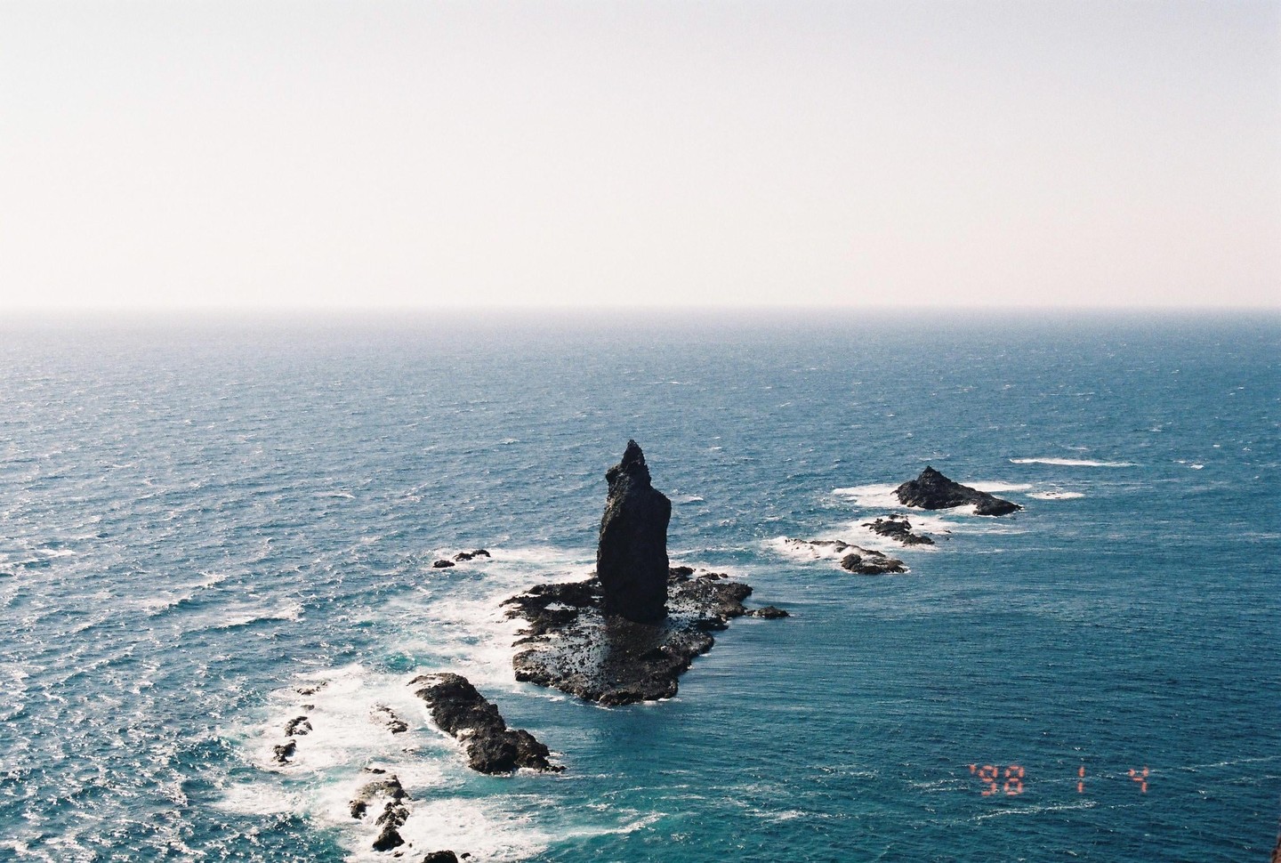 A tall, dark rock formation stands in the middle of a vast blue ocean with smaller rocks surrounding it.