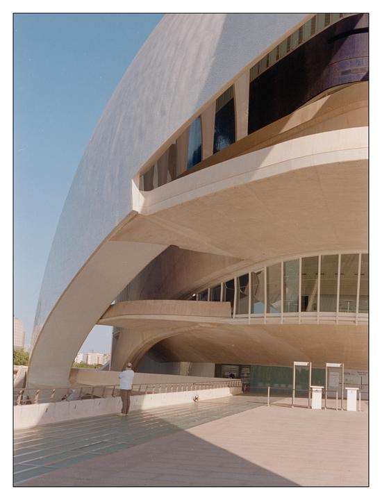 A man stands on a walkway next to a large, curved, futuristic building under a clear sky.