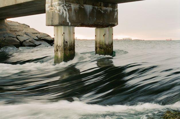 The tide rushes around the concrete pillars of a bridge, with rocks and a distant ship visible under a cloudy sky.