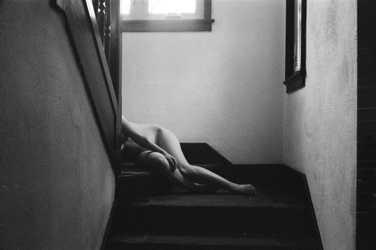 A nude woman is lying on a carpeted staircase next to a window.