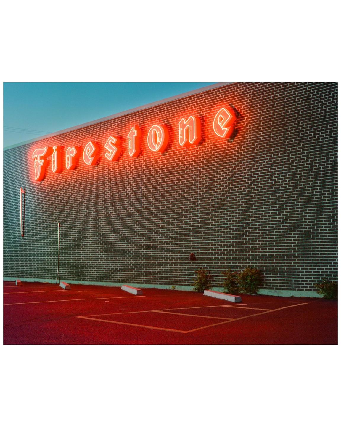 A neon "Firestone" sign glows red above a parking lot at dusk.
