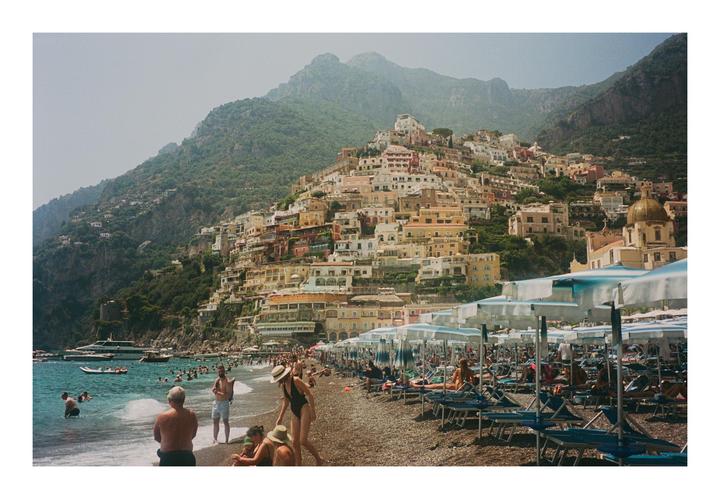 A crowded beach with many people and umbrellas is in the foreground, with a town built into a steep hillside in the background.