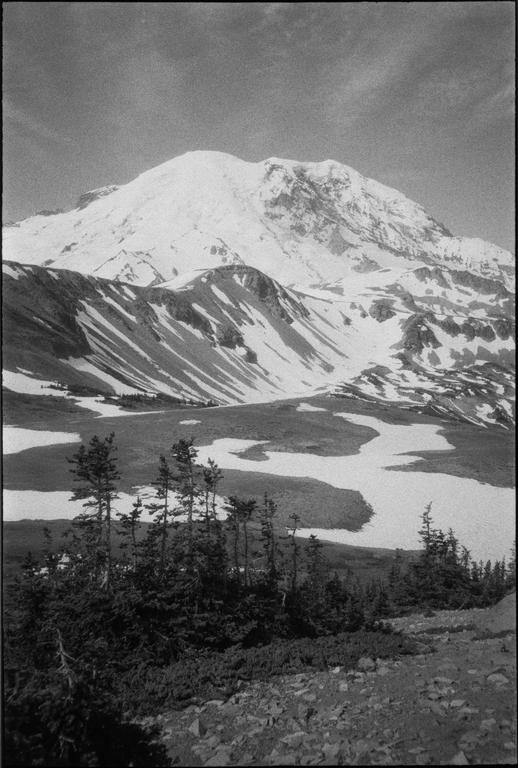 A snow-capped mountain rises above a valley with patches of snow and evergreen trees.