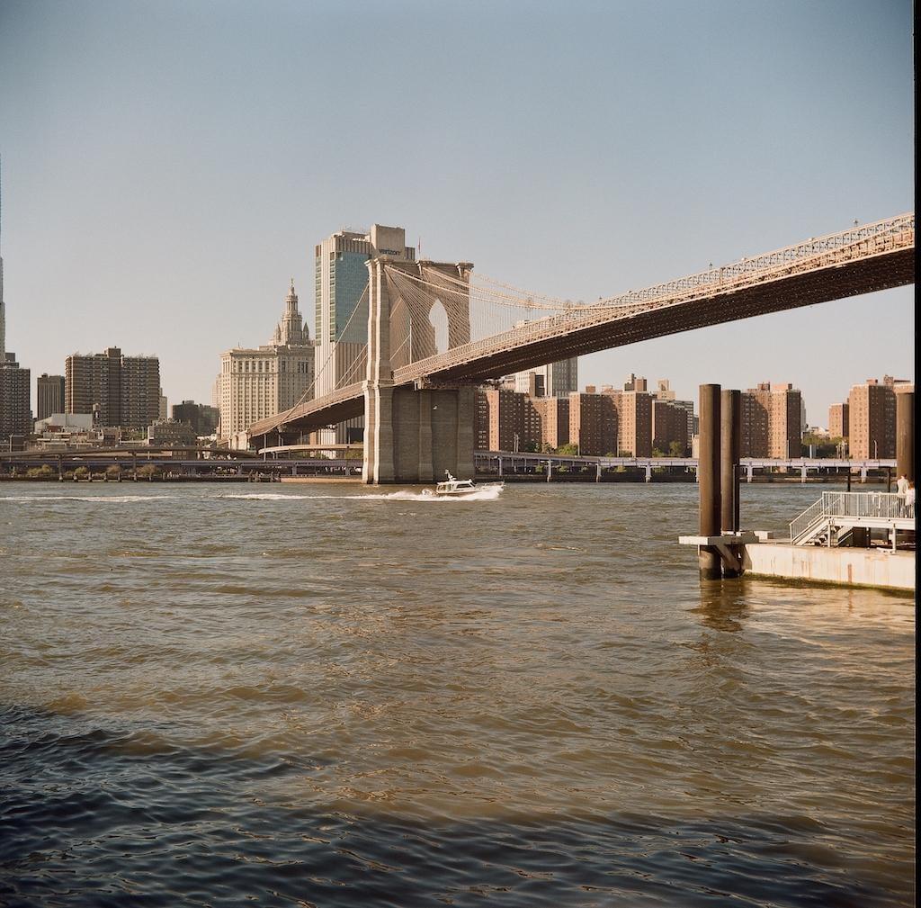 A boat moves across the water with the Brooklyn Bridge and city buildings in the background.
