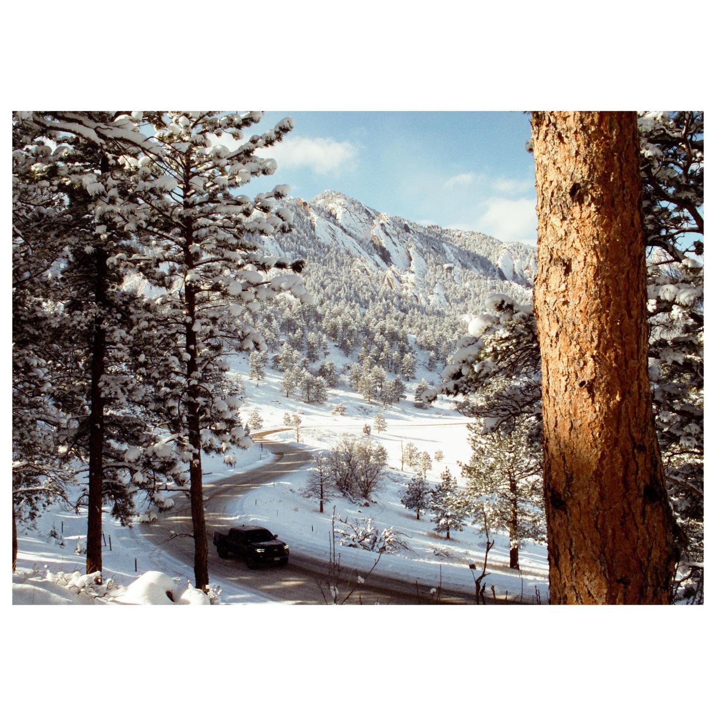 A winding road leads through a snow-covered forest towards a mountain range under a blue sky.