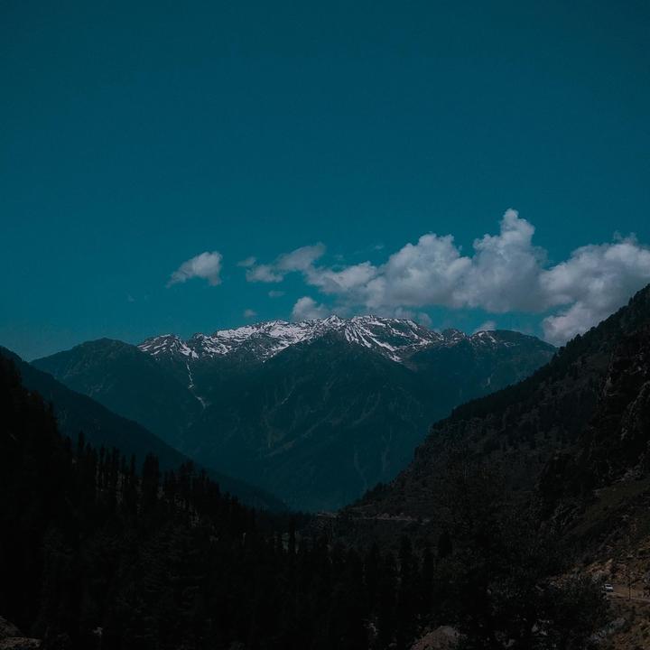 Snow-capped mountains rise above a valley with dark trees under a blue sky with white clouds.