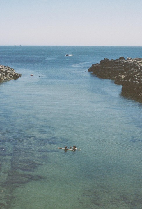 Two people swim in a clear blue cove with rocky shores under a bright sky.