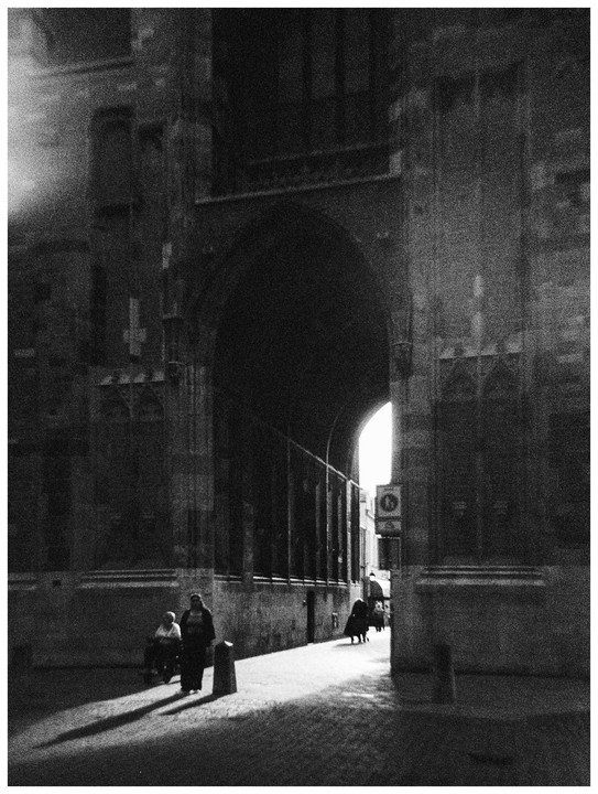 Two women stand in a sunlit archway while other people walk further down the street.