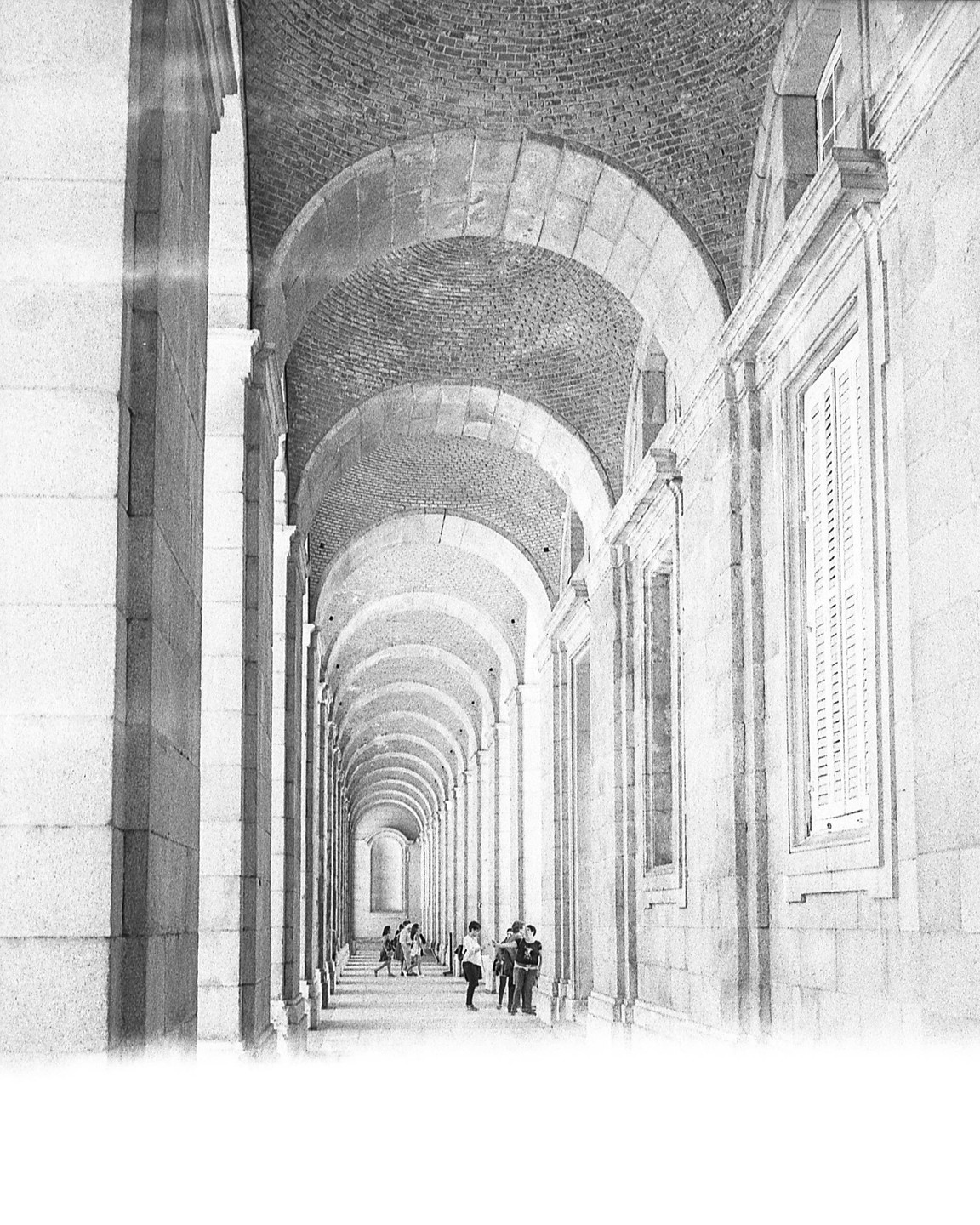 A long, arched hallway with brick ceilings and stone columns leads to a distant light, with several people walking and standing along the path.