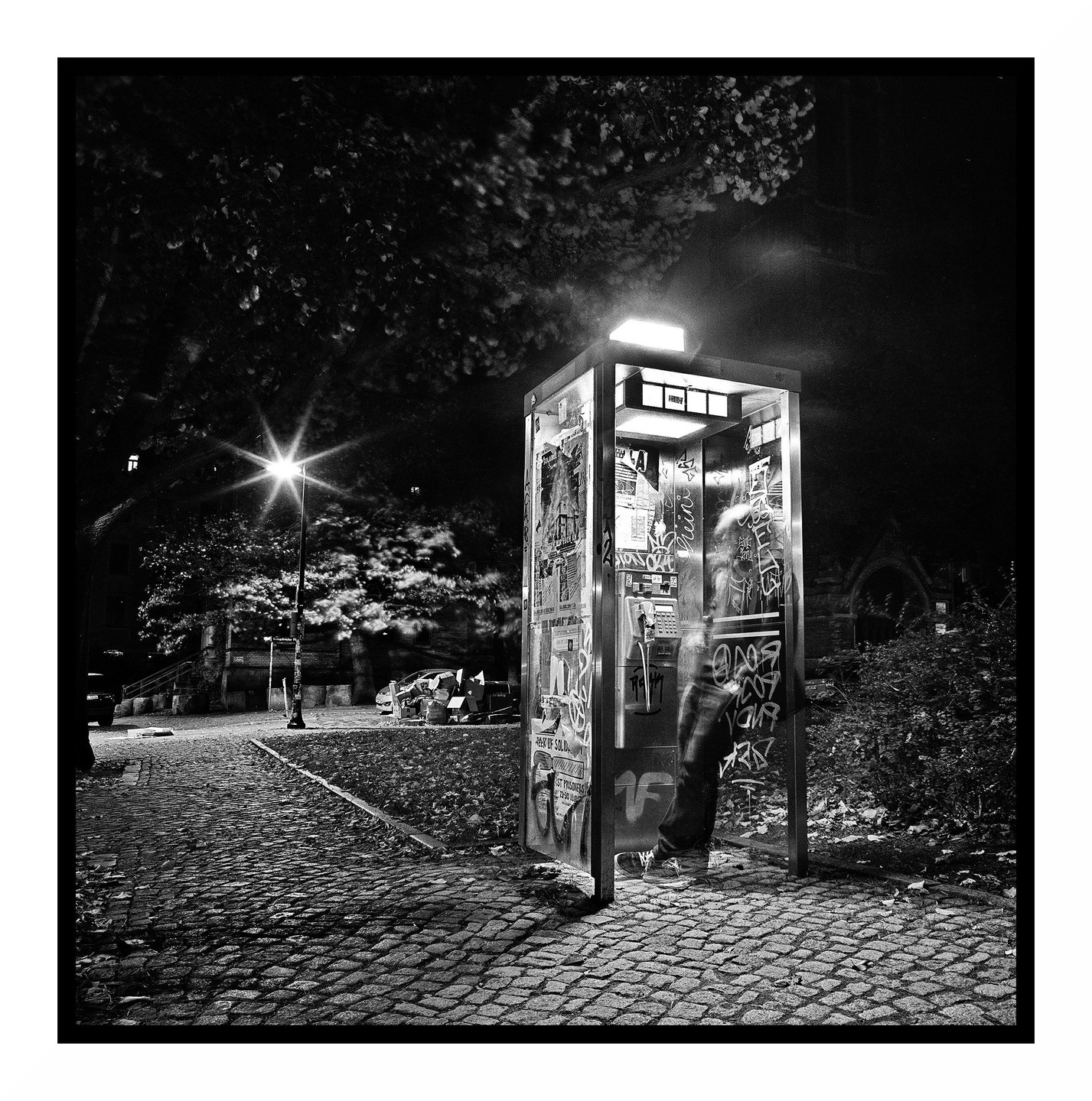 A person stands inside a graffiti-covered telephone booth on a cobblestone street at night.