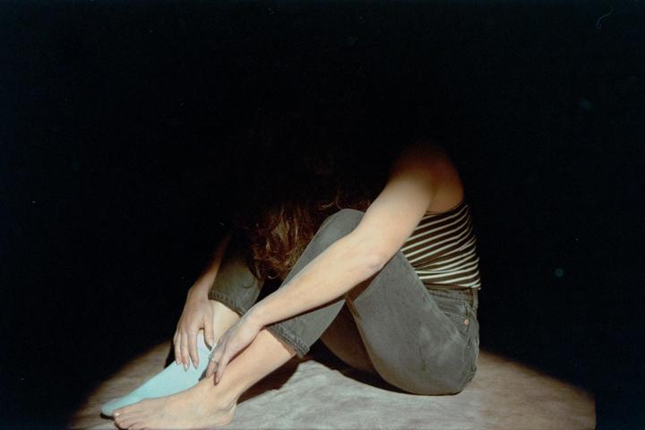 A woman sits on the floor with her head down, illuminated by a single spotlight.