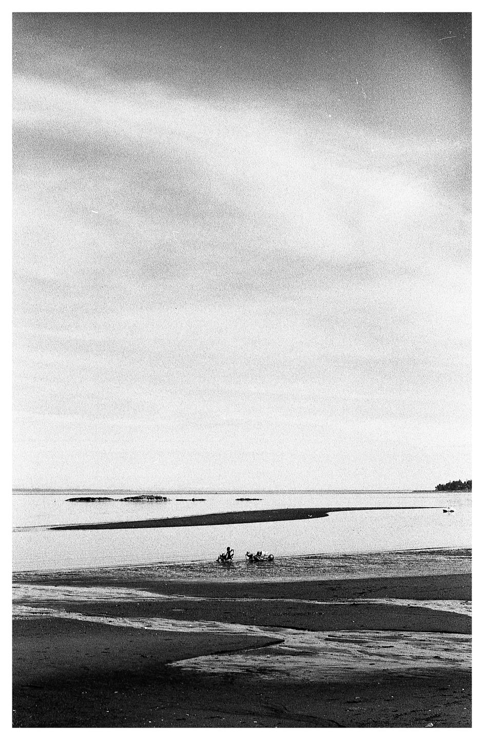 Two people are sitting in shallow water on a beach with a wide expanse of sky above them.