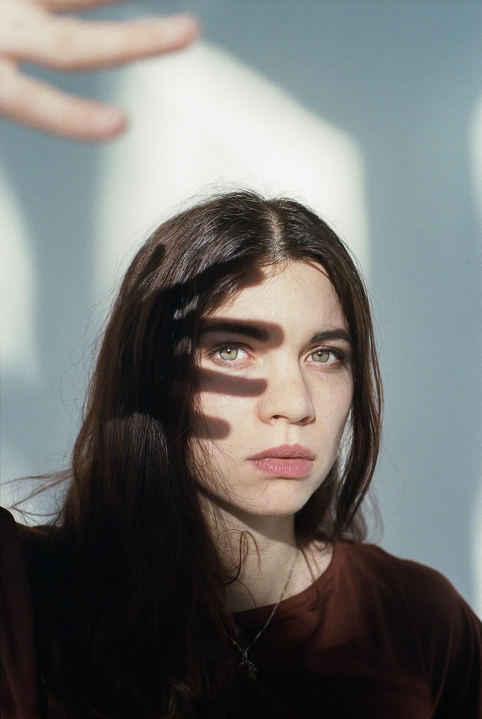 A woman with long dark hair looks directly forward, with a hand casting a shadow over her face.