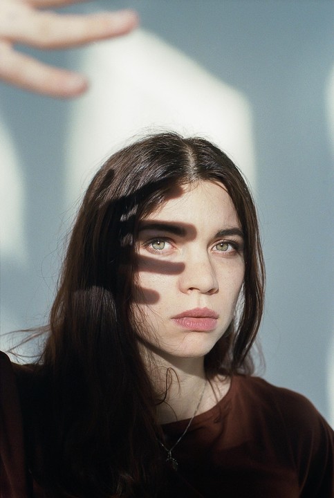 A woman with long dark hair looks directly forward, with a hand casting a shadow over her face.