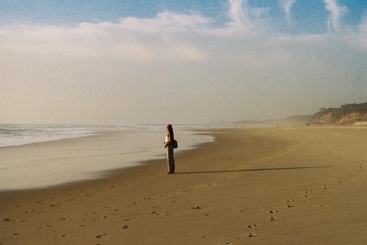 A woman stands on a sandy beach looking out at the ocean waves under a hazy sky.