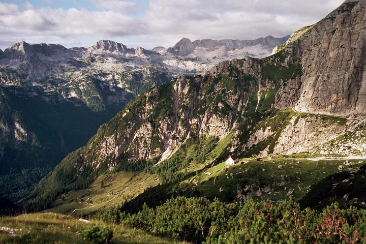 A small white house sits on a grassy mountain slope with a winding road and towering peaks in the background.