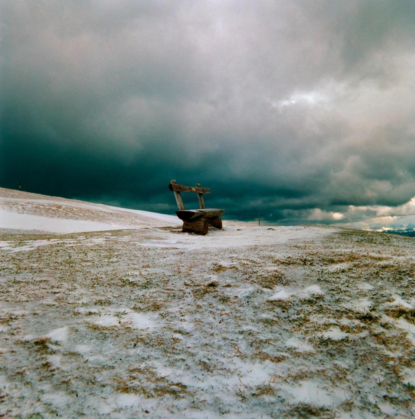 A wooden bench sits on a snow-dusted hill under a dramatic, cloudy sky.