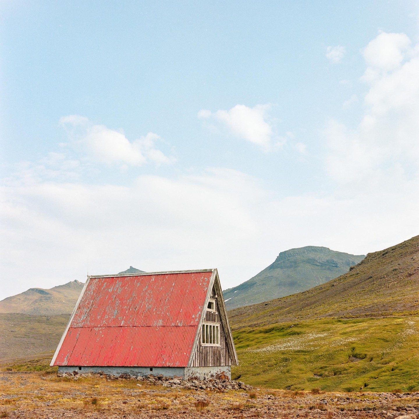 An A-frame cabin with a red roof sits in a vast, rocky landscape with mountains in the background.